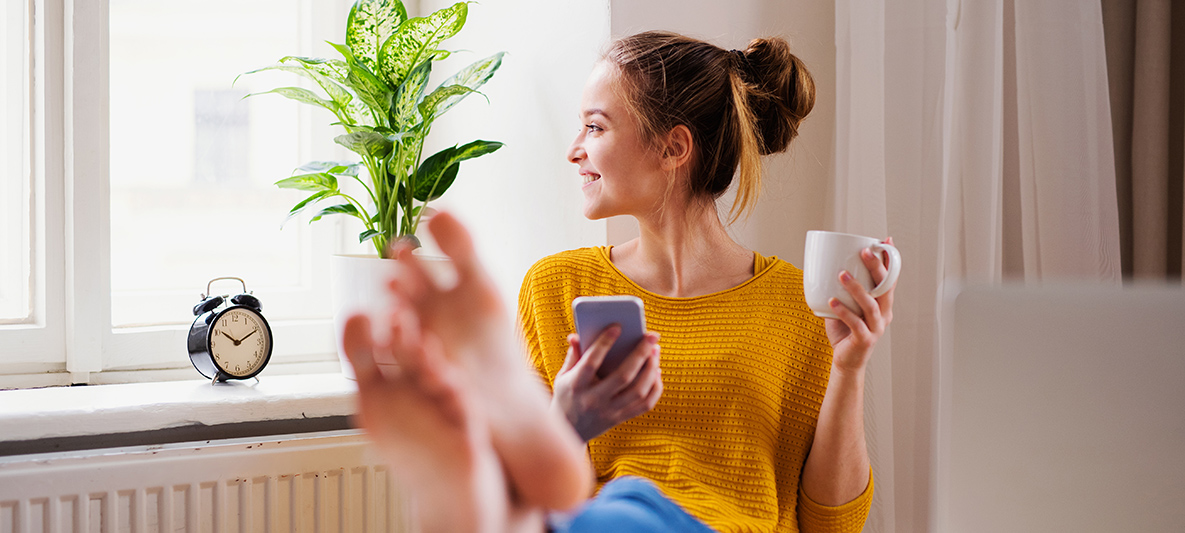 Frau sitzt mit Kaffee in ihrer Wohnung und guckt lächelnd aus dem Fenster Frau sitzt mit Kaffee in ihrer Wohnung und guckt lächelnd aus dem Fenster