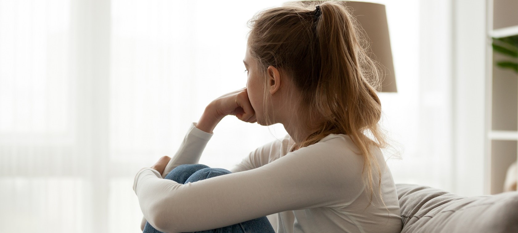Junge Frau sieht nachdenklich aus Fenster und hält Beine umschlungen Junge Frau sieht nachdenklich aus Fenster und hält Beine umschlungen
